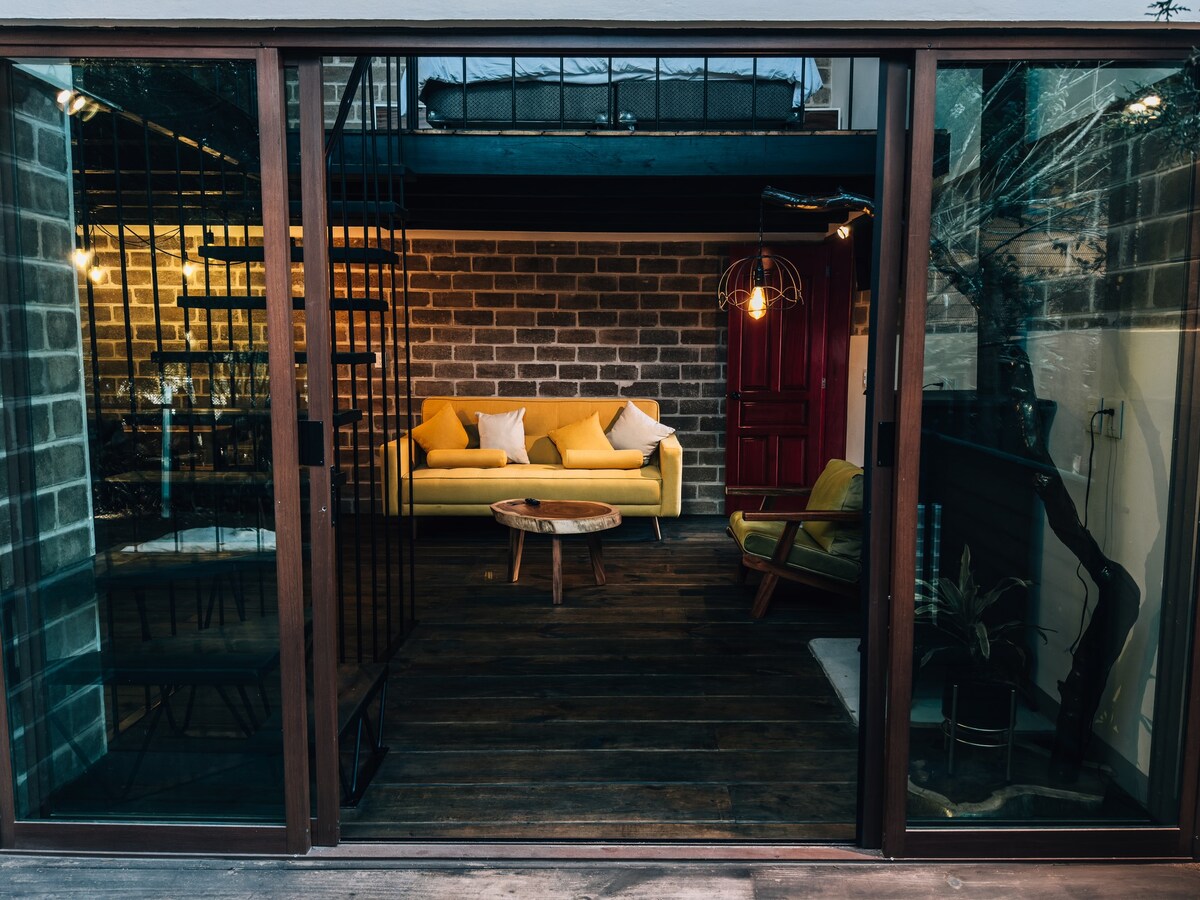 A modern living area is framed by large glass doors, featuring a bright yellow sofa paired with comfortable armchairs. A wooden coffee table sits in front, while a rustic stone wall adds texture to the space. Warm light from the overhead fixtures provides a cozy ambiance.