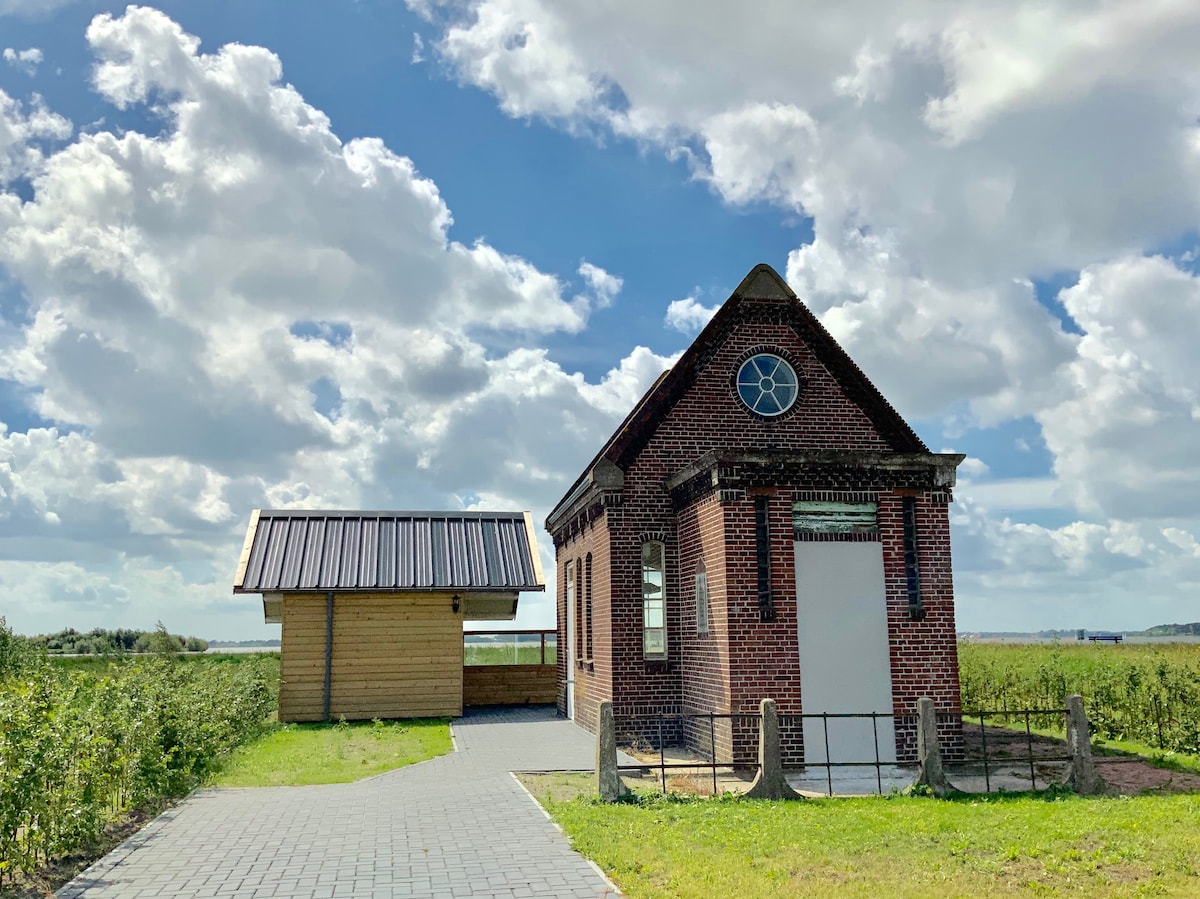 A restored historic building is positioned next to a modern wooden structure. The brick exterior features a prominent circular window and decorative details. A paved path leads toward the entrance, surrounded by grassy areas and open fields, under a sky adorned with fluffy clouds.