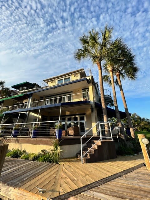 The three-story condo is situated by the water, featuring a modern architecture style with large balconies. Tall palm trees frame the exterior, and a wooden deck leads to the entrance. Clear skies and scattered clouds provide a serene backdrop to the property.