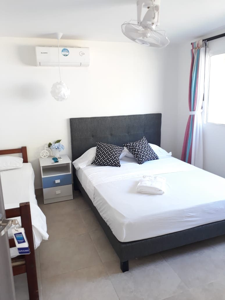 A well-lit sleeping area features a spacious bed with decorative pillows, alongside a smaller single bed. A nightstand with a lamp and a vase of flowers adds a touch of decor. Air conditioning and bright curtains enhance the comfort of the room.