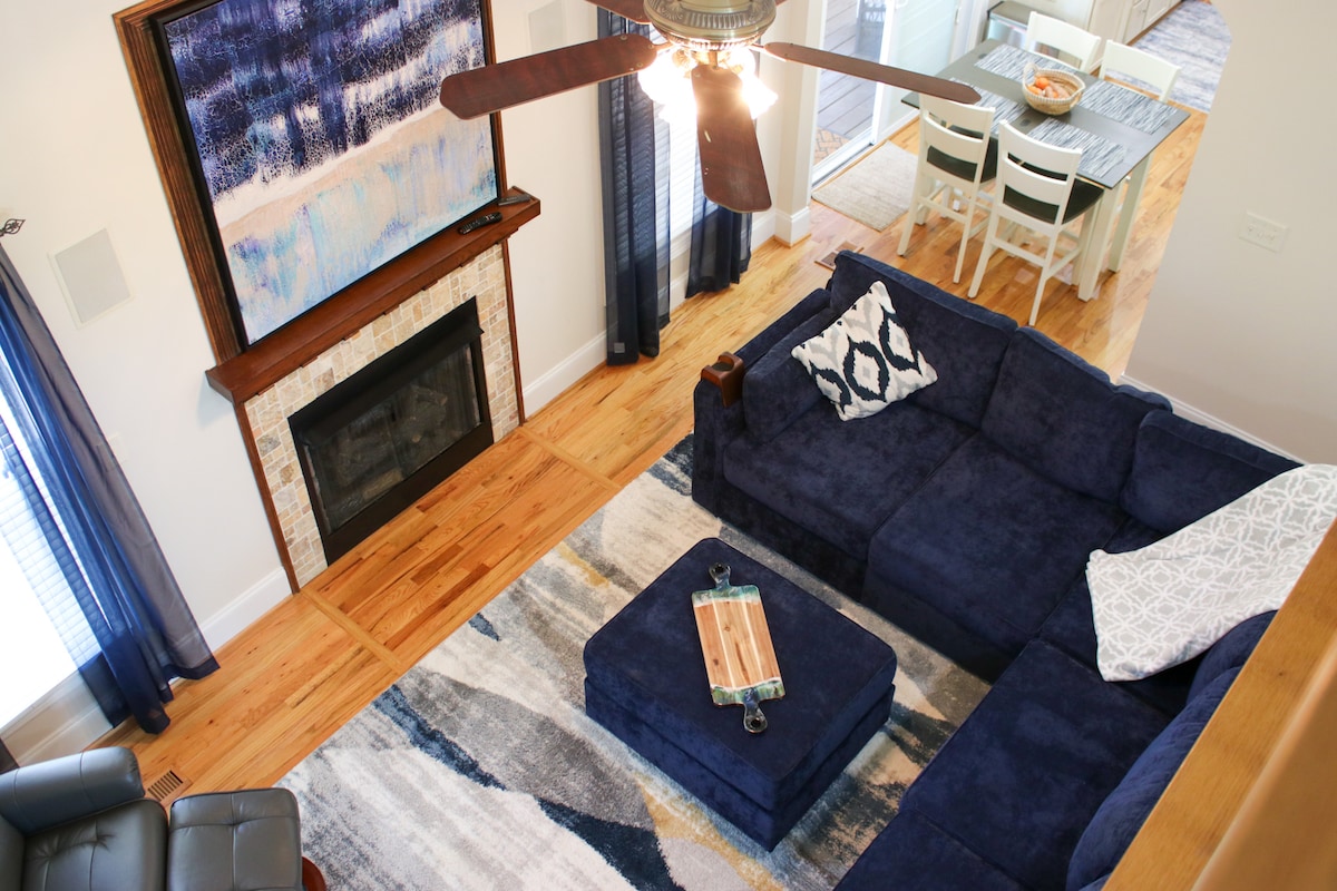 A living area is featured with a blue sectional sofa and coordinating ottoman. A textured area rug offers a soft touch underfoot. Natural light enters through large windows, while a modern fireplace is framed by a wooden mantel. A dining area can be seen in the background.