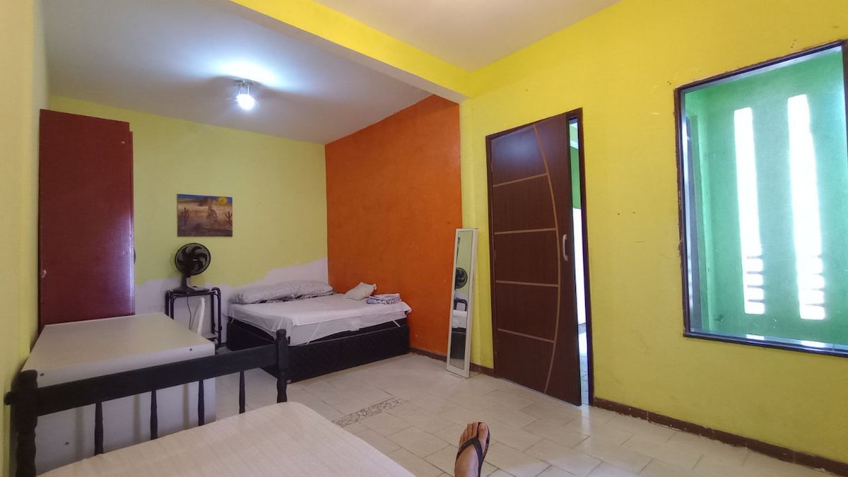 A vibrant room features a mix of yellow and orange walls. Two beds are positioned on either side, with a mirror standing in the corner. A fan is visible near the window, allowing for ventilation. Natural light enters through the green window nearby.
