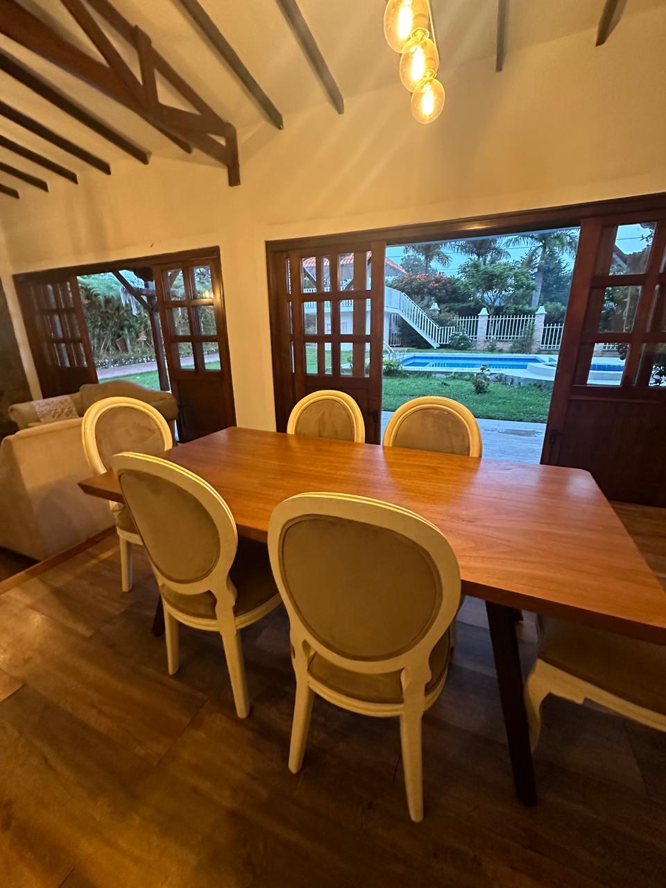 A wooden dining table is surrounded by four upholstered chairs, positioned near large windows that offer views of the outdoor greenery. Natural light filters in, illuminating the space. The ceiling features exposed beams, enhancing the rustic character of the room.