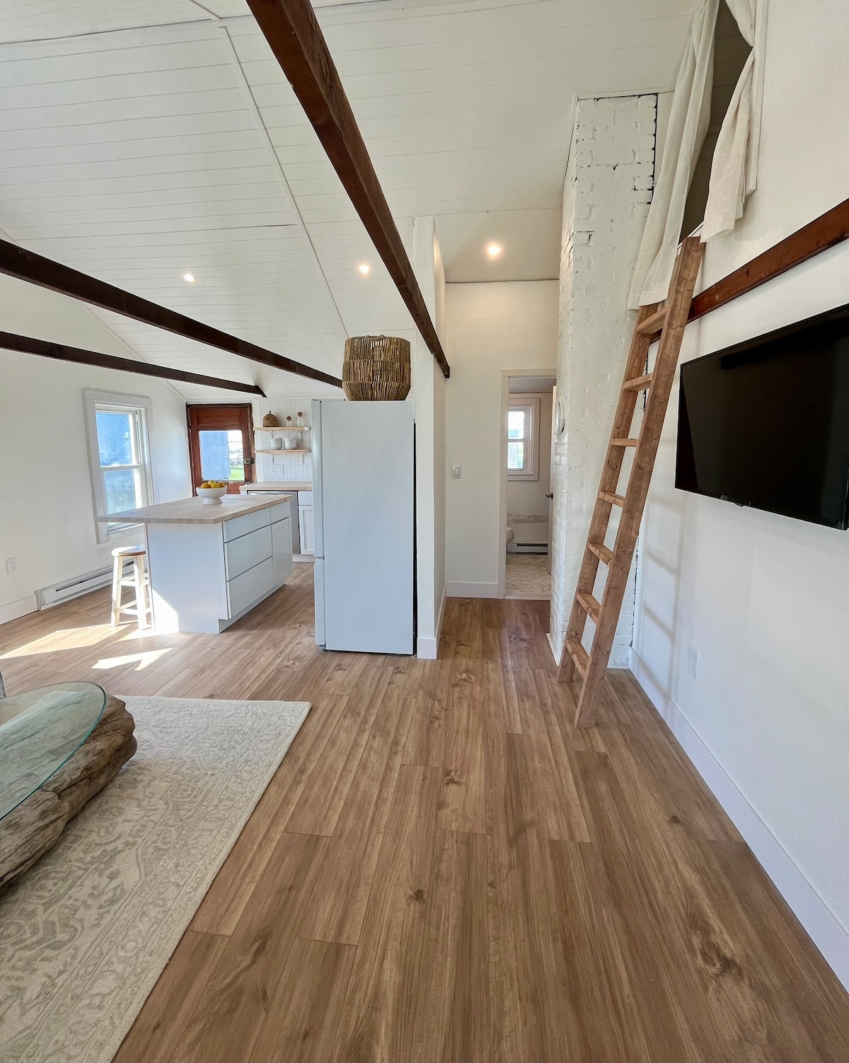 An open living area features a wooden ceiling with exposed beams. Light wood flooring extends throughout, complemented by a light rug. A kitchen area is visible in the background, with white cabinetry and a bar counter with seating. A wooden ladder leads to the loft space.