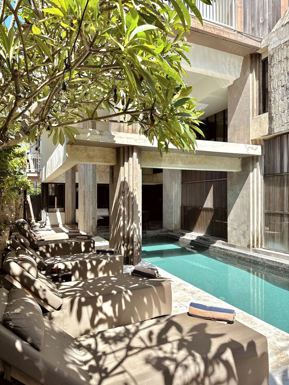 A tranquil outdoor space is presented, featuring a sleek swimming pool bordered by comfortable lounge chairs. Sunlight filters through the leaves of a nearby tree, casting playful shadows across the patio area. The modern architecture is complemented by natural stone elements, creating a serene atmosphere.