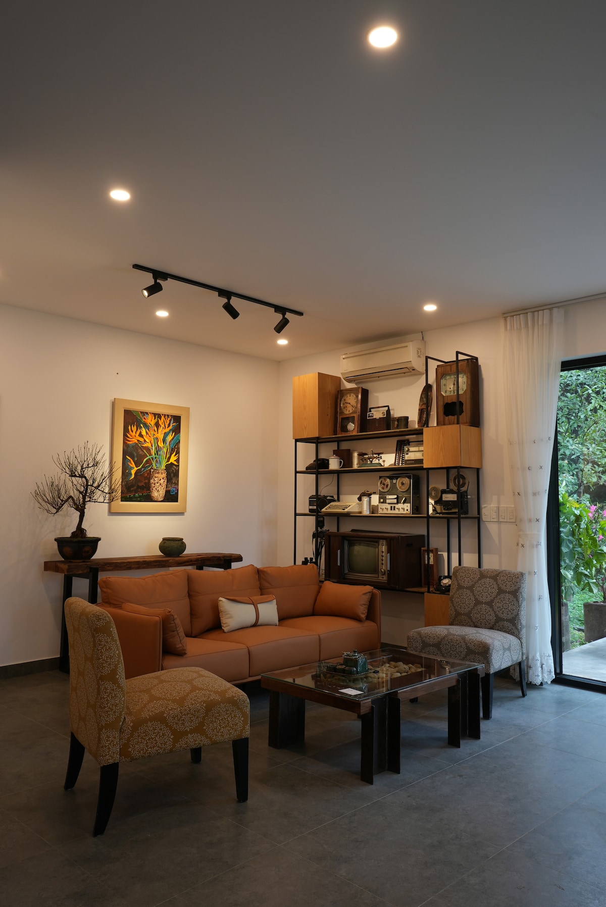 A modern living space features an orange sofa paired with patterned chairs, arranged around a glass-top coffee table. Decorative elements, including a wall-mounted art piece and plants, enhance the ambiance, while natural light filters in through large windows.
