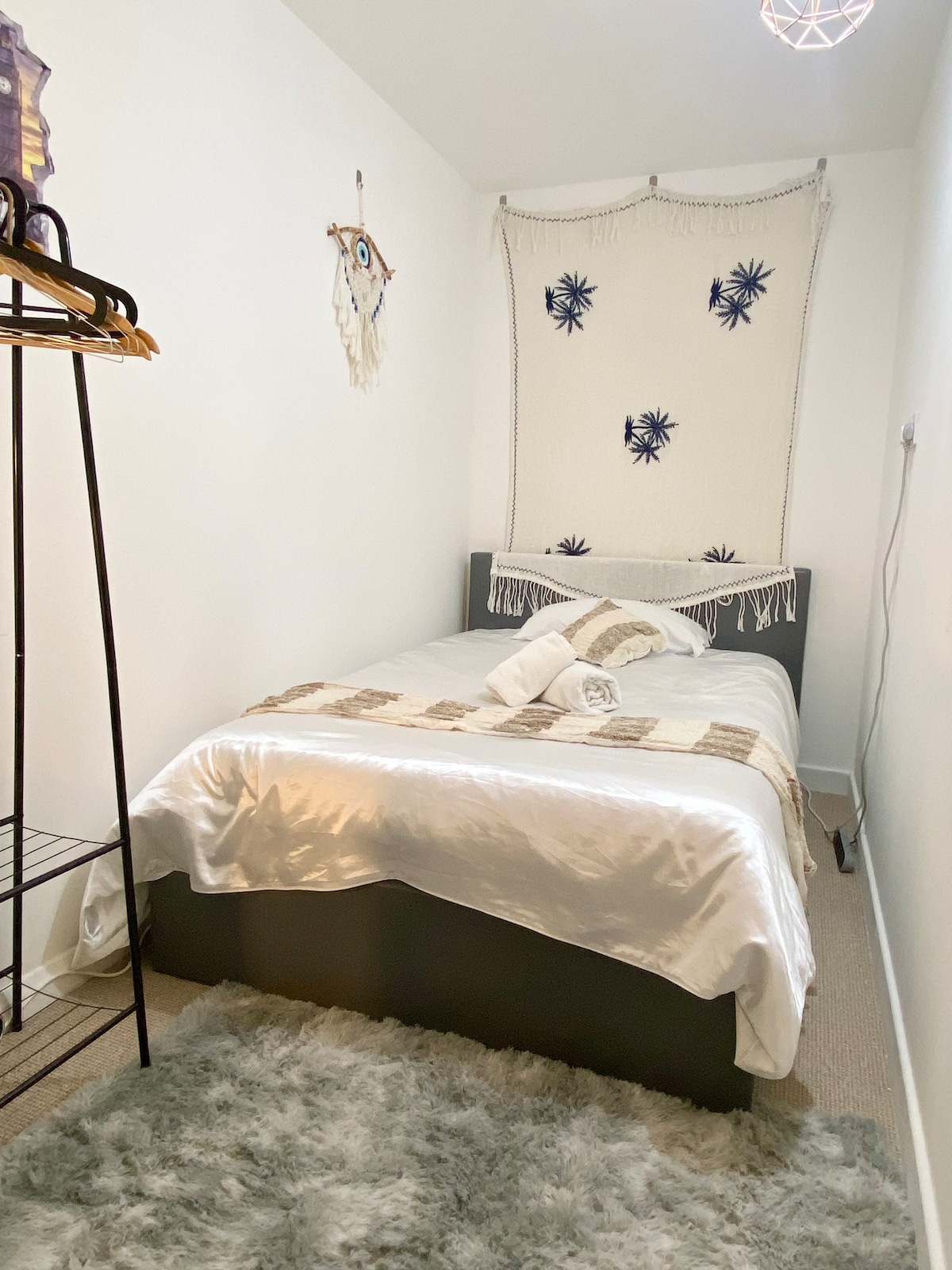 A cozy bedroom is furnished with a double bed, topped with a light, decorative blanket and neatly rolled towels. A soft rug covers the floor, while wall hangings and a clothes hangar enhance the bohemian decor. Natural light subtly illuminates the space.