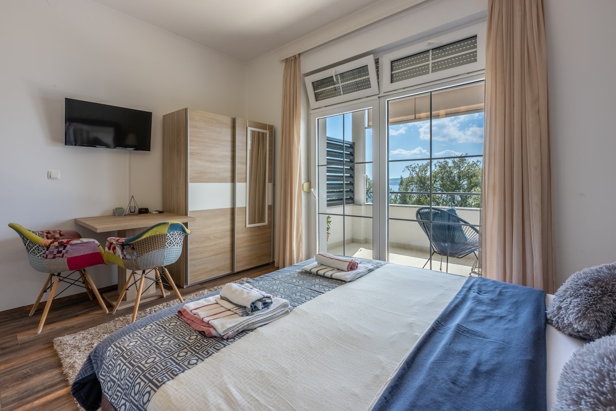 A spacious sleeping area features a large bed with neatly arranged linens. A wooden side table and a television mount are positioned nearby. A small desk with colorful chairs faces a large window, allowing natural light to fill the room and providing views of the outdoors.