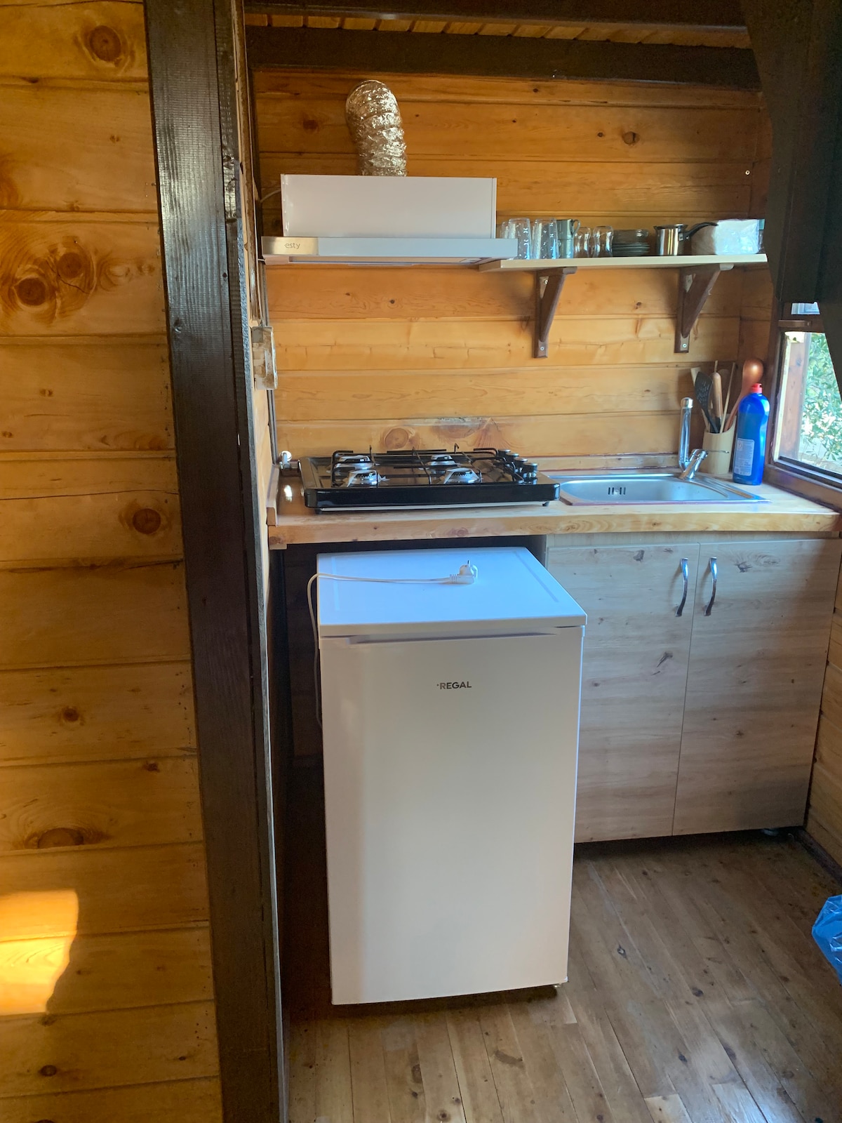A compact kitchen area features wooden walls and a light wood countertop. A small white refrigerator is prominently displayed next to a gas stove, with a sink and shelves holding various kitchen items visible above. Natural light filters in through a nearby window.