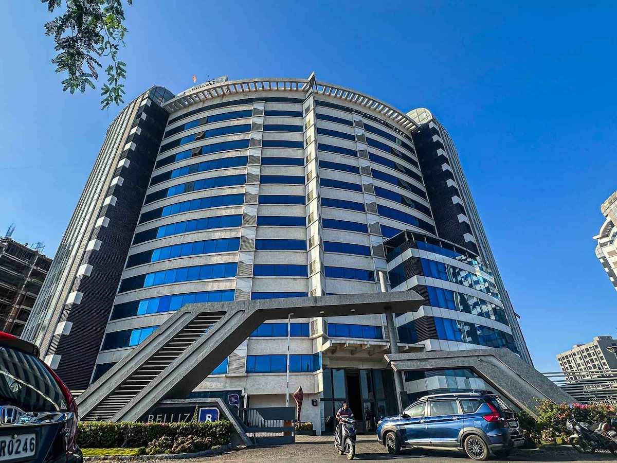 A modern multi-story building features a sleek façade of glass and dark steel. Large windows reflect the clear blue sky, while bold architectural lines draw the eye towards the entrance. Vehicles are parked in the foreground, providing context for the urban environment.