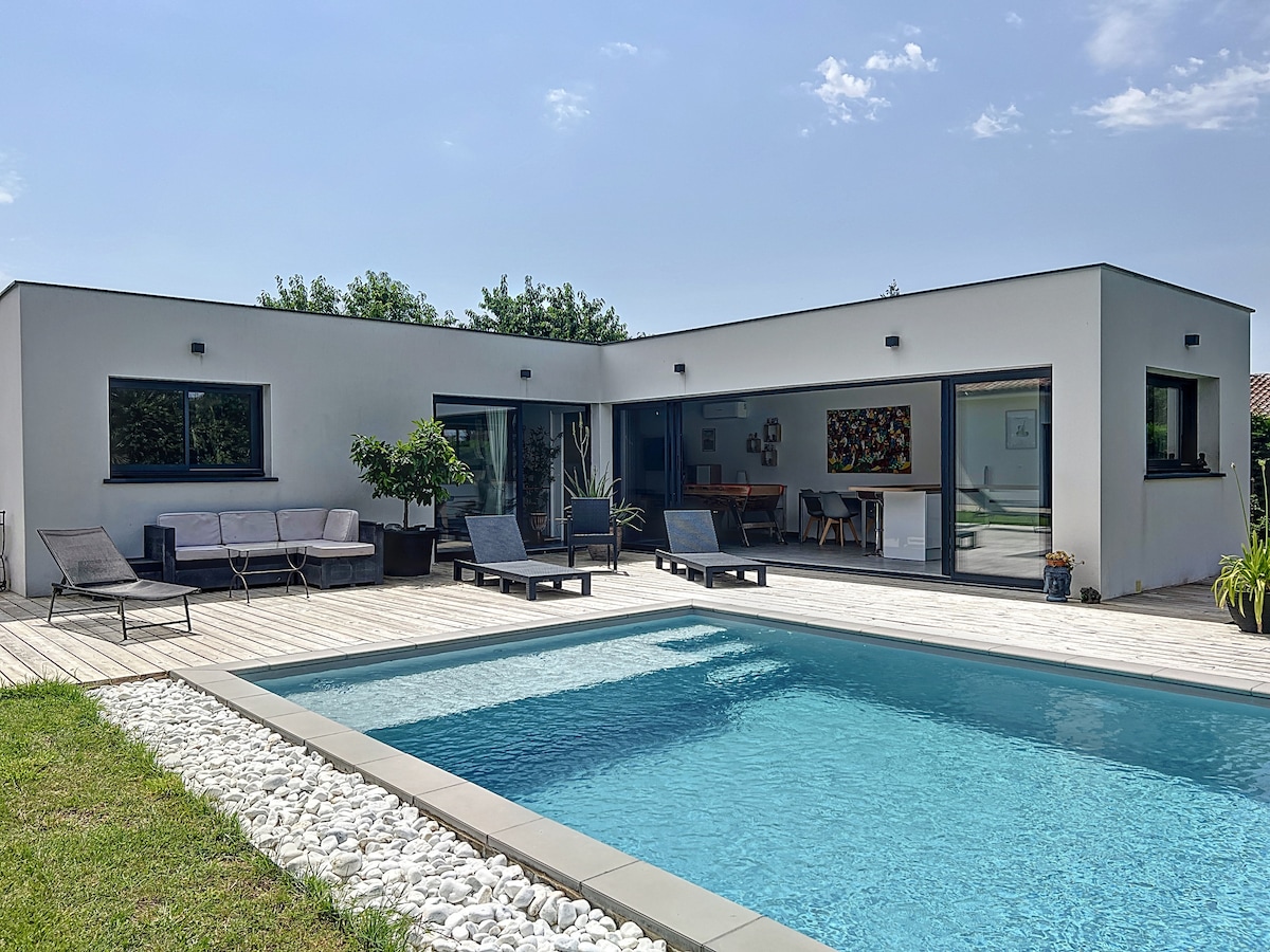 An independent apartment is showcased in this image, featuring a swimming pool surrounded by a wooden deck. Comfortable lounge chairs are positioned near the pool. Large windows and sliding doors enhance natural light access, revealing a cozy living area visible through the glass.