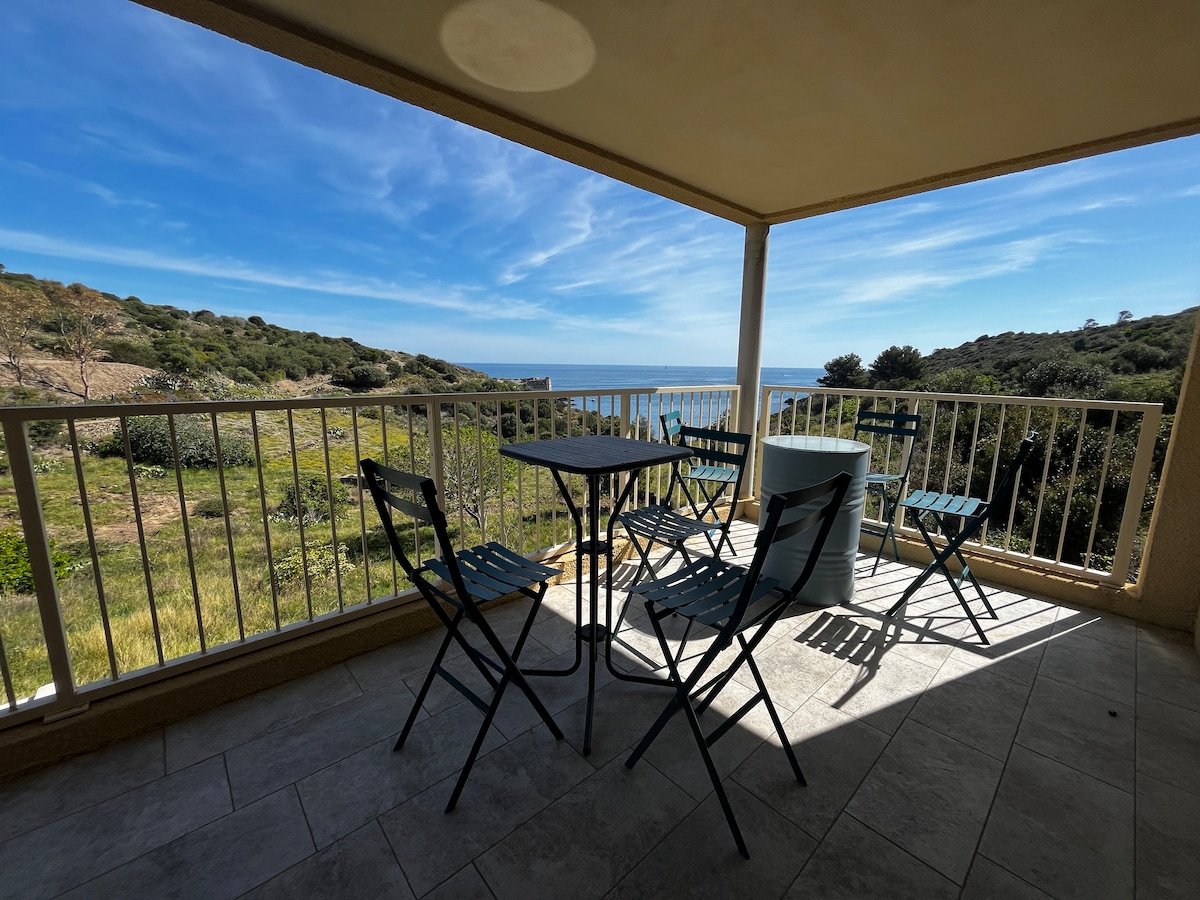 A private terrace is shown, featuring a small round table surrounded by four folding chairs. The landscape includes a view of rolling greenery and the sea beyond, with blue skies and wispy clouds enhancing the serene setting.