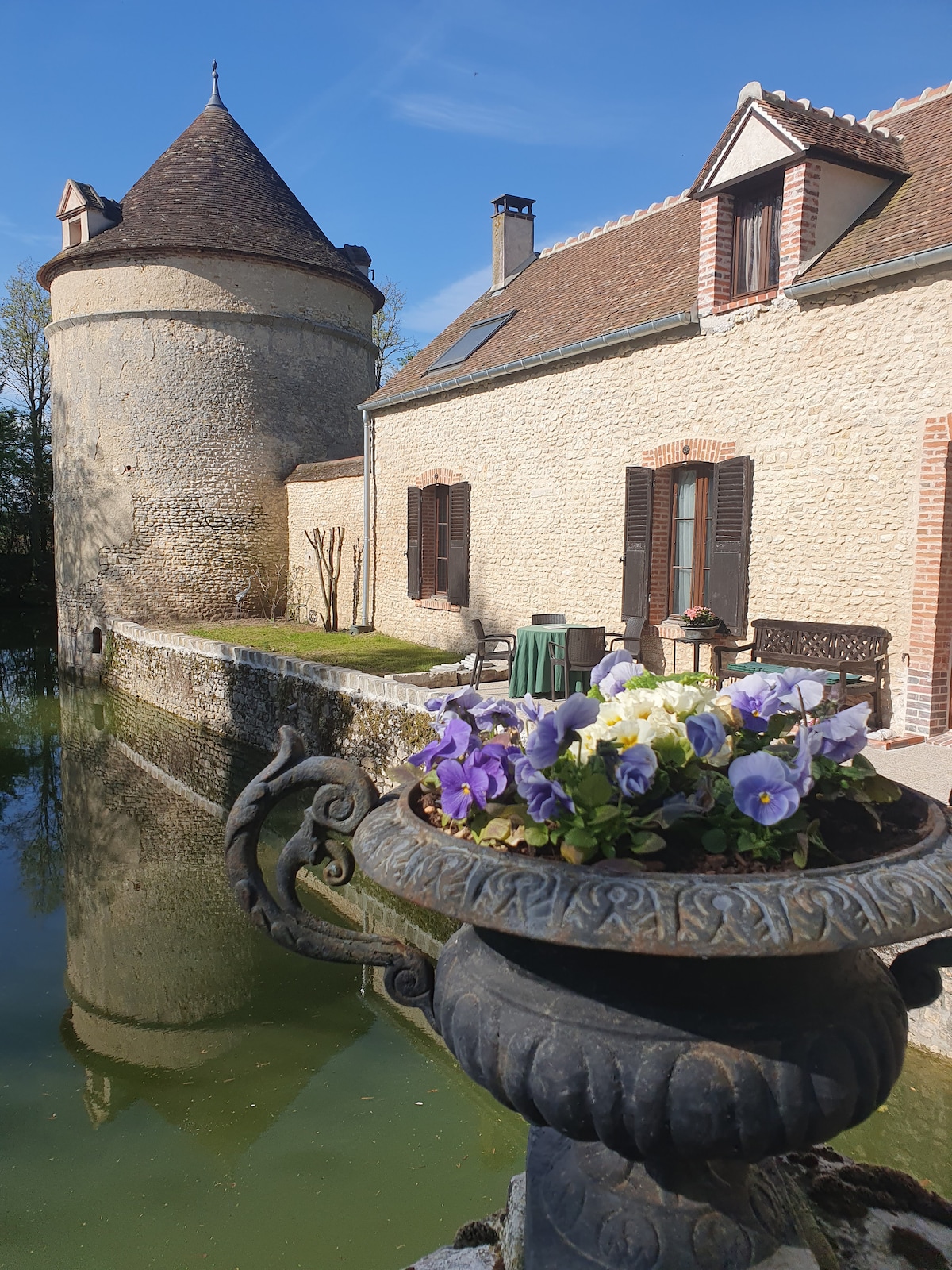 An outdoor scene captures a stone tower beside a tranquil pond. In the foreground, an ornamental urn is filled with vibrant purple and yellow flowers. The building's textured stone walls and wooden shutters create a peaceful ambiance, enhanced by the clear blue sky.
