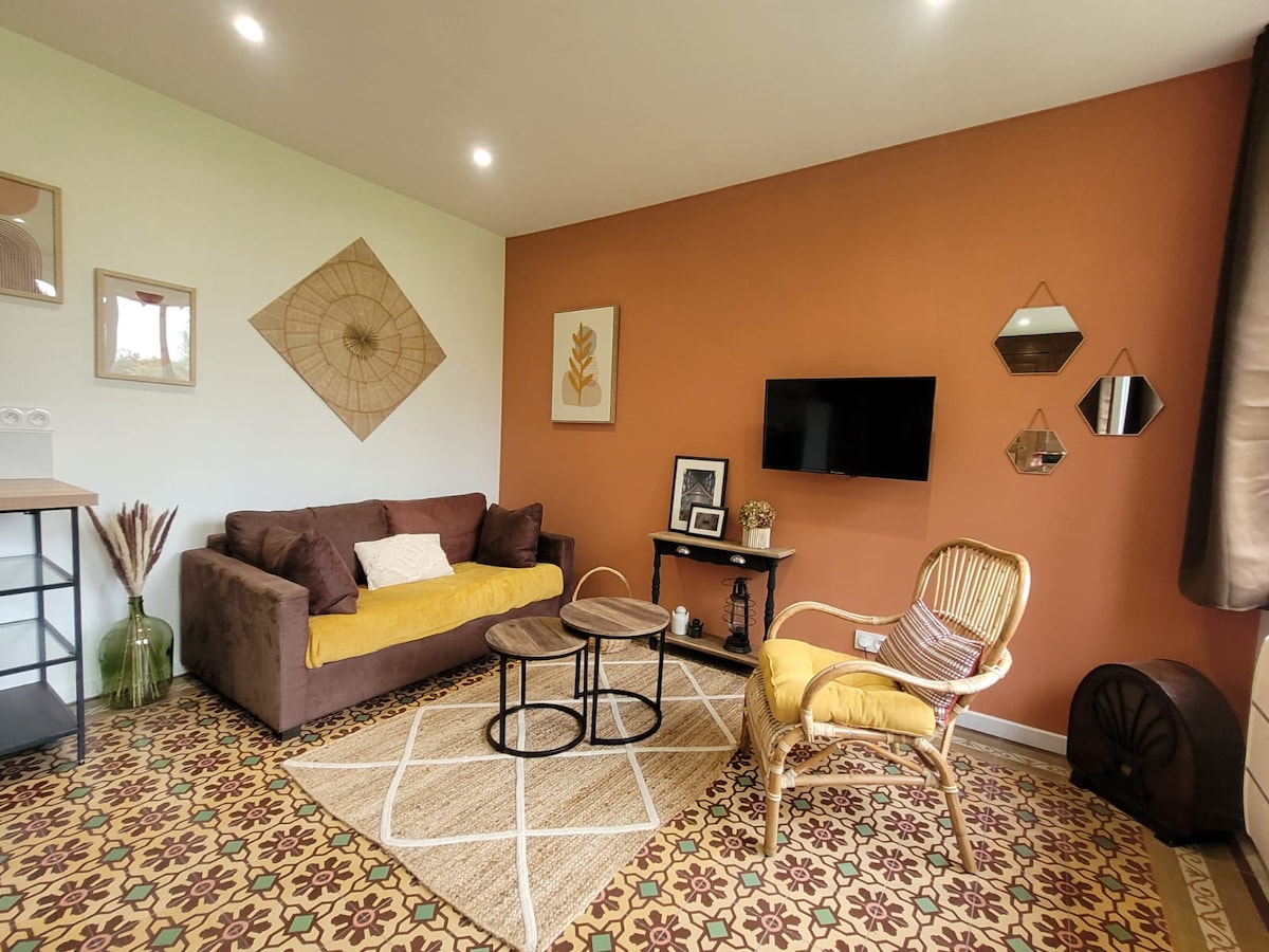 A cozy living area features a dark brown couch with yellow accents, complemented by a wicker chair. Two coffee tables sit on a patterned rug, while a flat-screen TV is mounted on the wall. Decorative wall art and soft lighting enhance the space.
