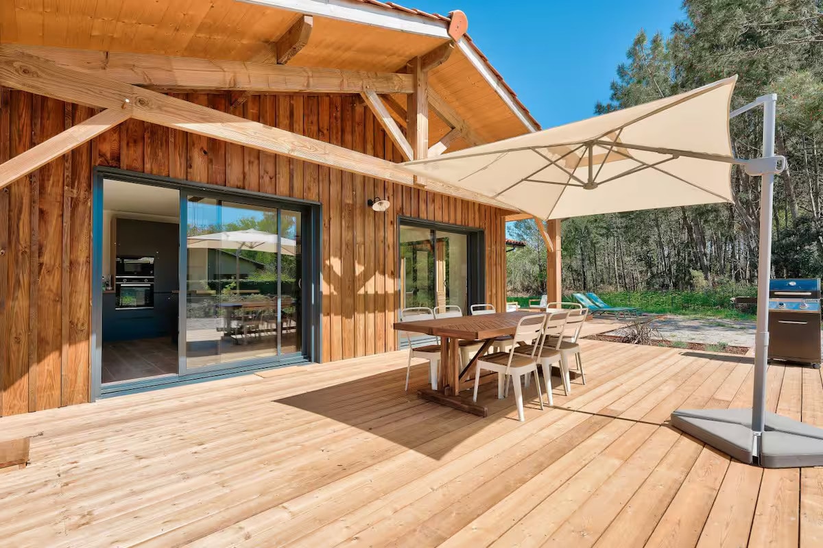 A spacious deck made of wooden planks features a large table surrounded by six chairs. A shade umbrella is positioned to the side. Glass doors lead into the interior, illuminating the area with natural light. Lush greenery is visible in the background.