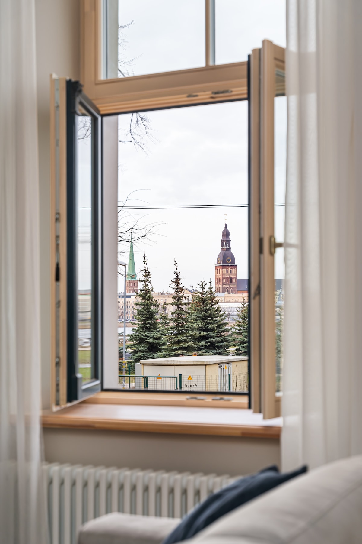A window is opened to reveal a scenic view of Riga's skyline, featuring a prominent tower and green trees in the foreground. Soft curtains frame the window, allowing natural light to illuminate the interior space.