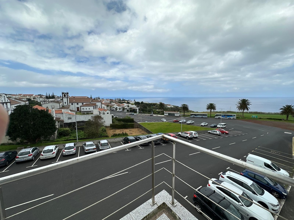 A view from a balcony overlooking a coastal neighborhood. The image captures a parking lot with several parked cars, while lush greenery and palm trees frame the seaside backdrop. In the distance, the ocean and a cloudy sky are visible, enhancing the serene ambiance of the area.