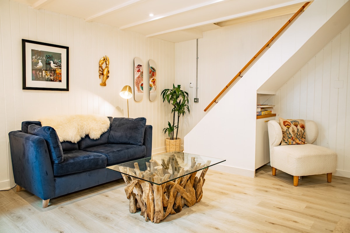 A comfortable living area features a navy blue sofa adorned with a textured throw, positioned next to a glass coffee table made from driftwood. Wall art adds character, while a potted plant and unique armchair create an inviting nook. Natural light brightens the space.