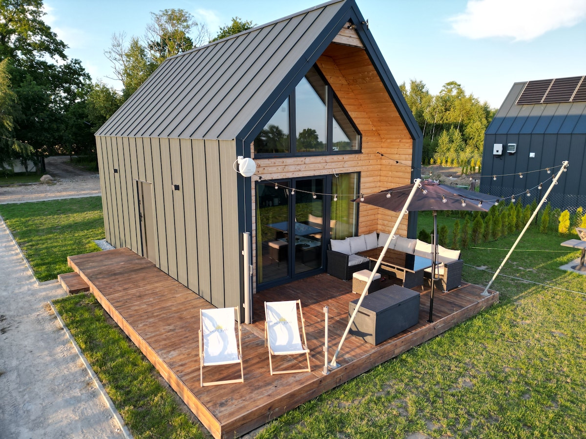 A modern barn-style house features a large deck with comfortable outdoor seating and an umbrella. The structure, with a mix of wood and metal, showcases stunning angular lines and expansive glass doors that connect the interior to the outdoor space.