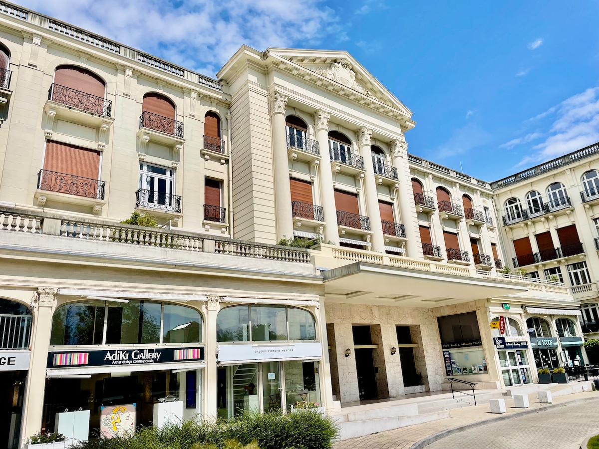 A historic building features elegant architectural details, including decorative columns and balconies. Large windows allow natural light to fill the space. The façade is complemented by a well-maintained entrance, surrounded by manicured landscaping, creating a welcoming atmosphere.