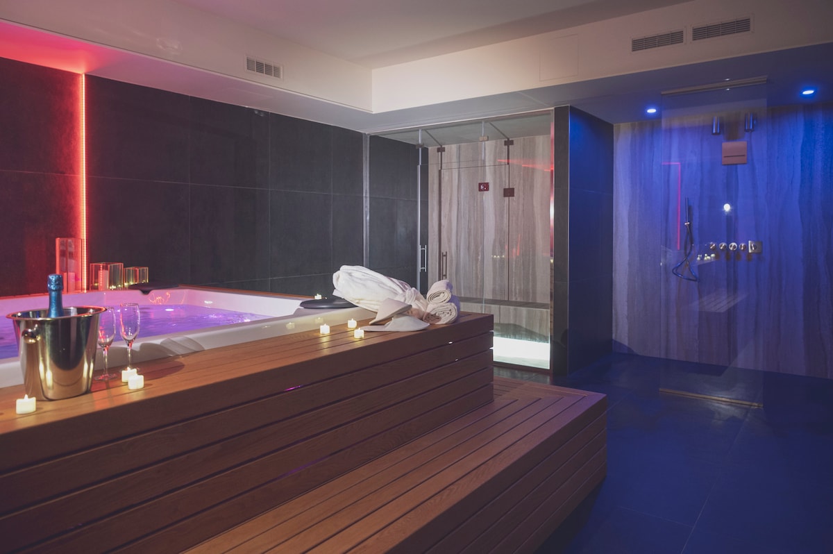 A private SPA area features a modern jacuzzi with a wooden platform surrounding it. Soft lighting casts a warm glow, while a steam room and shower are visible in the background. Candles are arranged on the surfaces, enhancing the tranquil atmosphere.