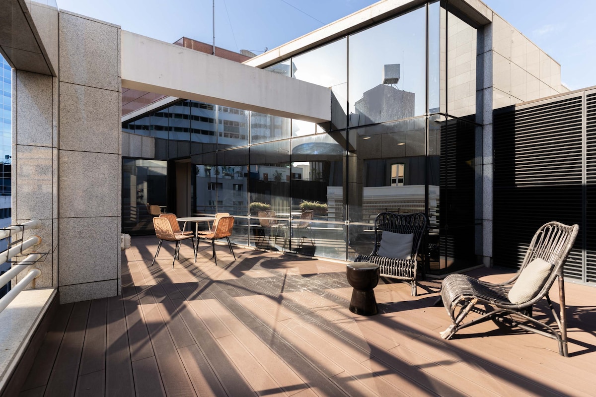 A spacious terrace features modern outdoor furniture, including a round table with four chairs and a lounge area with a cozy chair. Sunlight casts distinctive shadows on the wooden flooring, while the surrounding glass walls create a bright and open atmosphere.