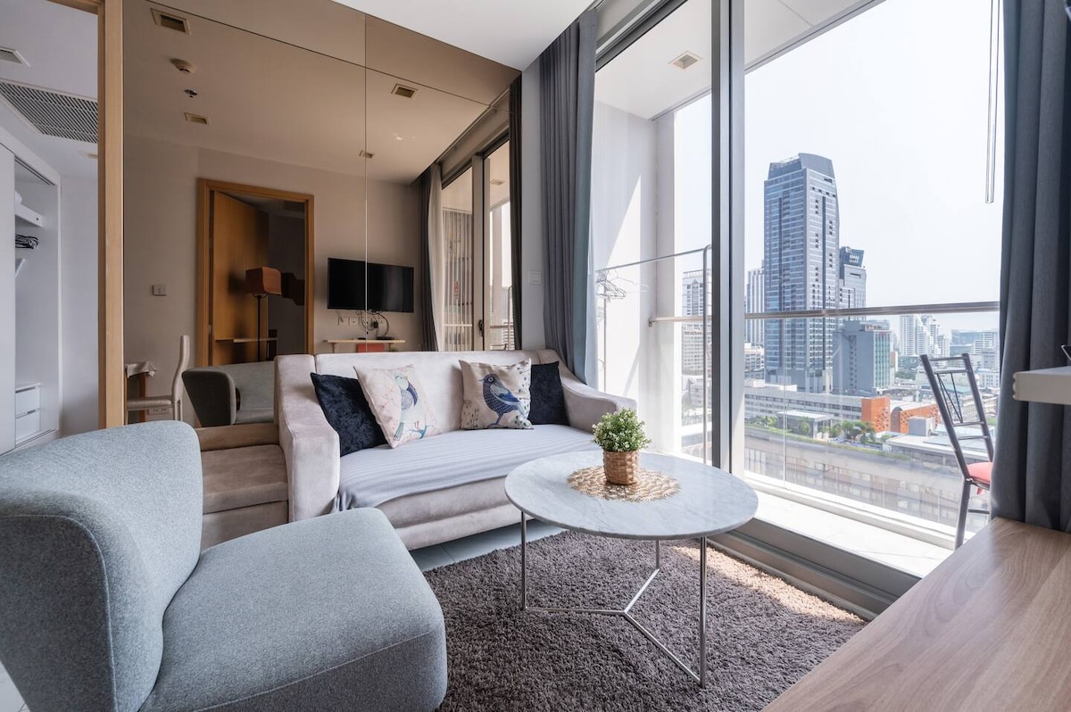 A living area features a light-colored sofa paired with decorative pillows, a round marble coffee table, and a plush rug. Large windows provide natural light and showcase a city view. A small dining table with chairs is visible in the background.