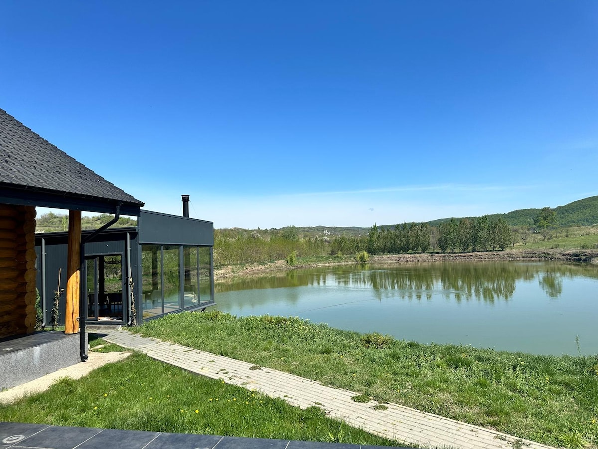 A serene view is presented, featuring a clear lake bordered by lush greenery. The dark wooden cabin sits on the left, while a spacious glass structure opens to the water. A stone path leads from the cabin to the lakeside, under a bright blue sky.