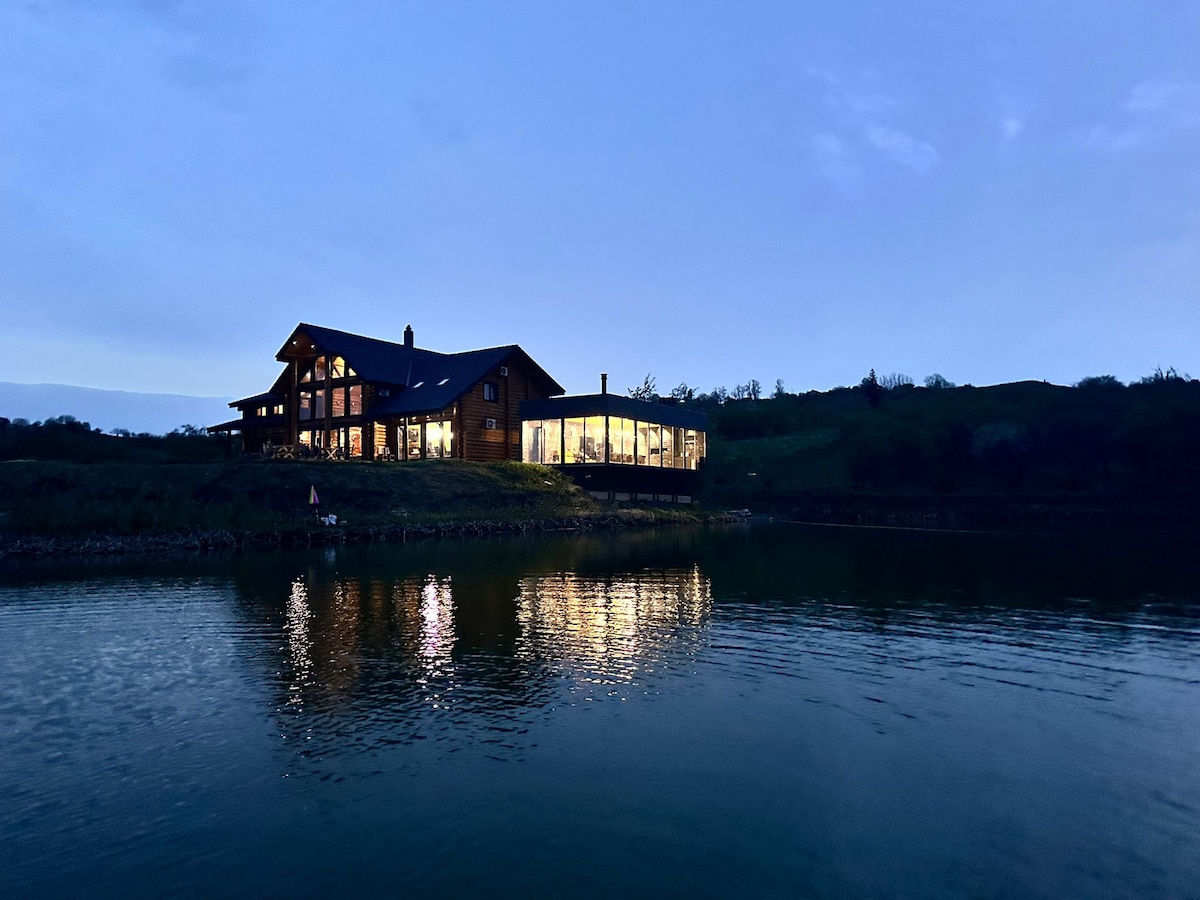 A wooden cabin is situated beside a calm lake, illuminated by soft lighting during twilight. The structure features large glass windows that reflect beautifully on the water's surface, while the surrounding landscape is softly outlined by the evening light.