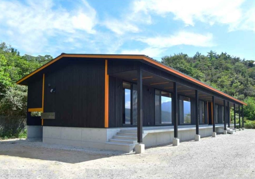 A modern, single-story villa is showcased, characterized by a dark exterior complemented by orange accents. Large windows allow natural light to fill the interior, while the surrounding greenery hints at a tranquil environment. Steps lead to the entryway and a gravel pathway frames the building.
