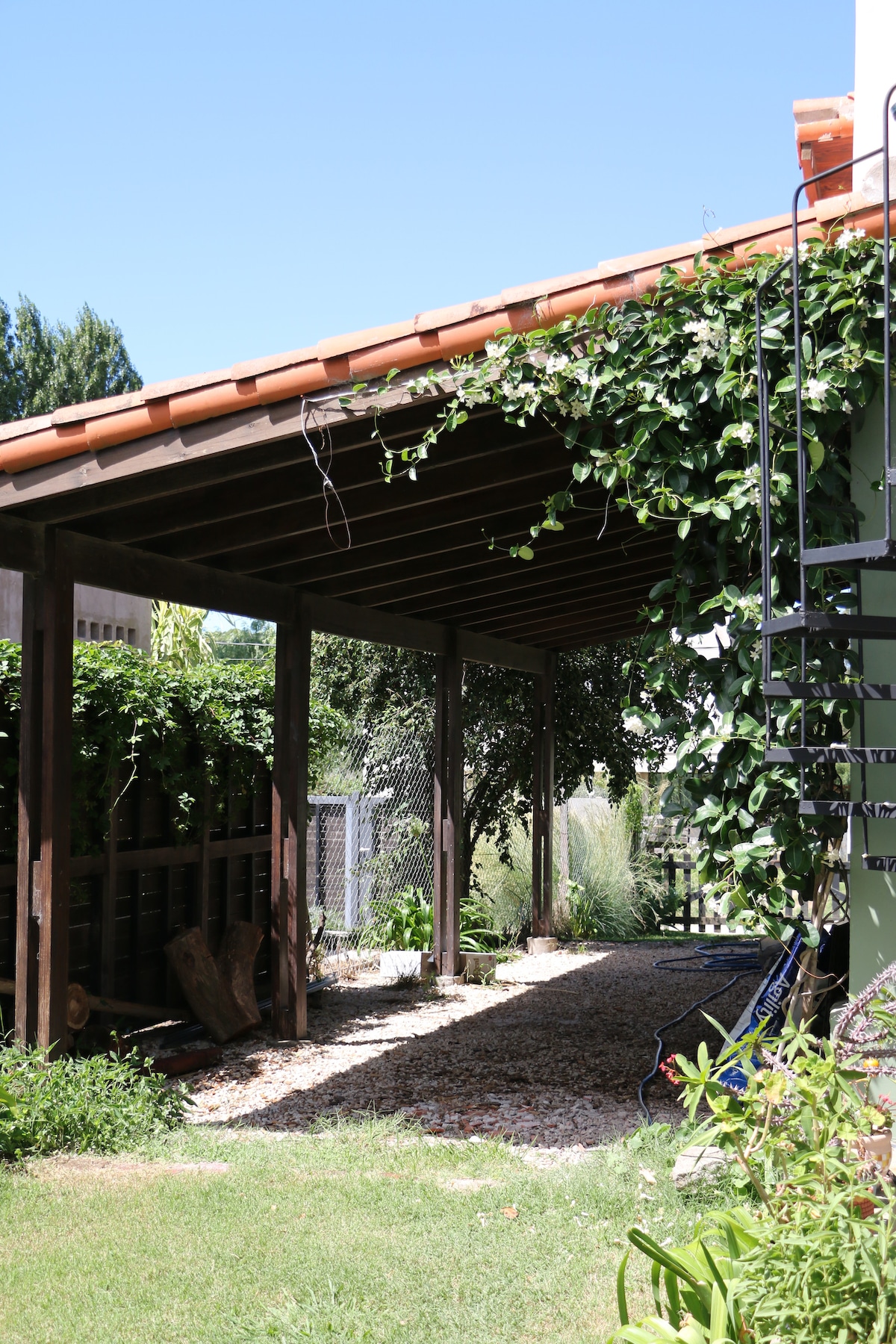 A covered outdoor area features a wooden structure adorned with climbing greenery. Gravel leads to a grass patch, creating a natural transition. The surrounding space includes plants, enhancing the serene atmosphere. A wrought-iron ladder is visible, offering access to the upper level.
