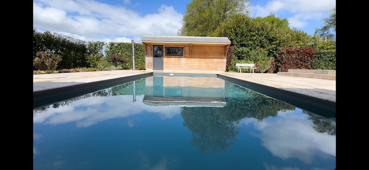 A serene outdoor pool area features a clear water surface reflecting the sky and surrounding greenery. A wooden structure is visible nearby, complemented by well-maintained landscaping. The tranquil setting invites relaxation, with ample space around the pool for lounging and enjoying nature.