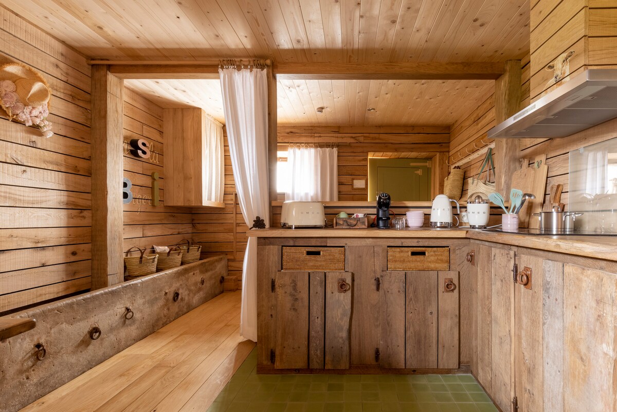 A rustic kitchen with wooden walls and flooring is featured, showcasing a range of white small appliances. A cozy dining area is visible through an opening, with soft natural light streaming through the curtains. Decorative baskets add a touch of warmth.