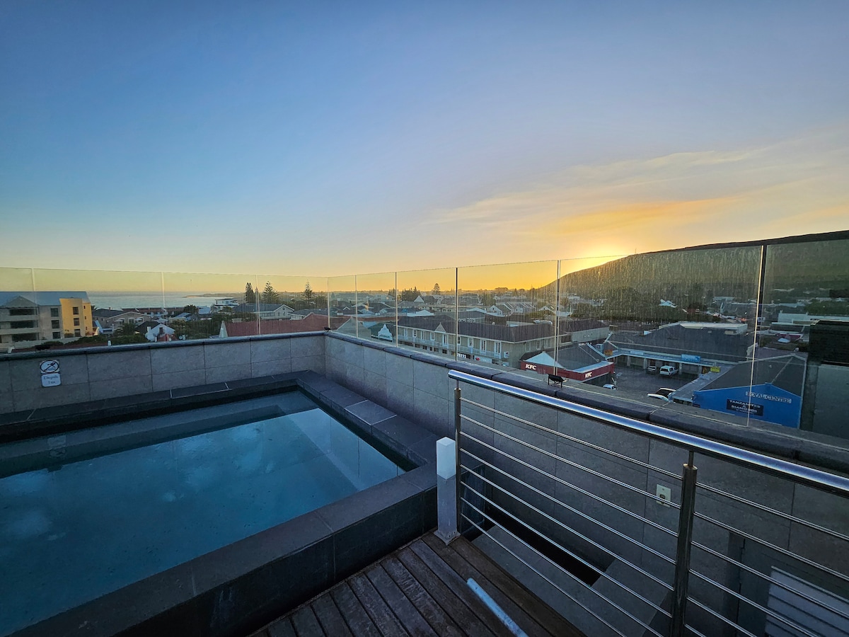 A rooftop pool is highlighted against a backdrop of a sunset over the town. Glass railings provide unobstructed views of the surrounding area, while the pool's serene surface reflects the evening light, creating a peaceful setting.