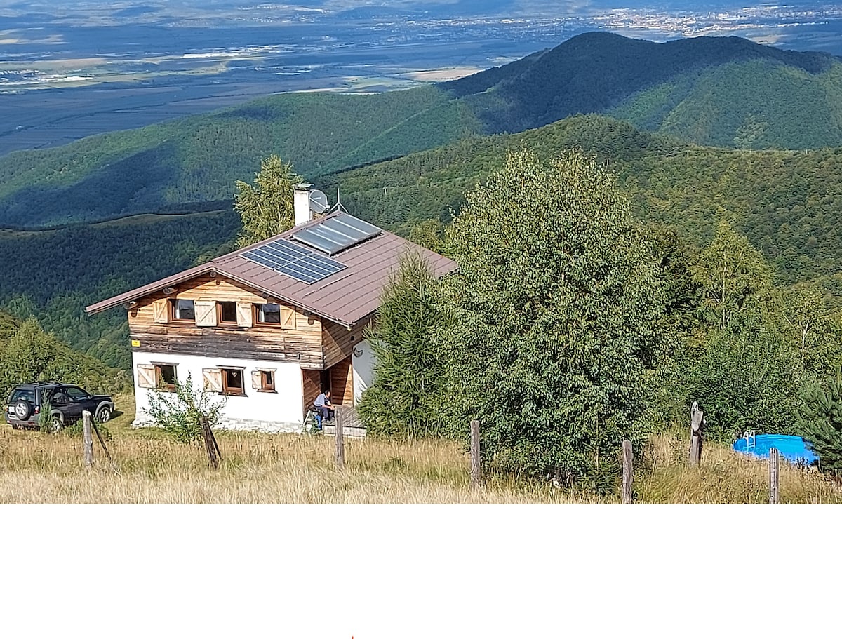 A spacious holiday home is nestled on a hillside, surrounded by lush greenery and rolling mountains. Solar panels are visible on the roof. A scenic view of the valley and distant hills enhances the serene atmosphere of the location.