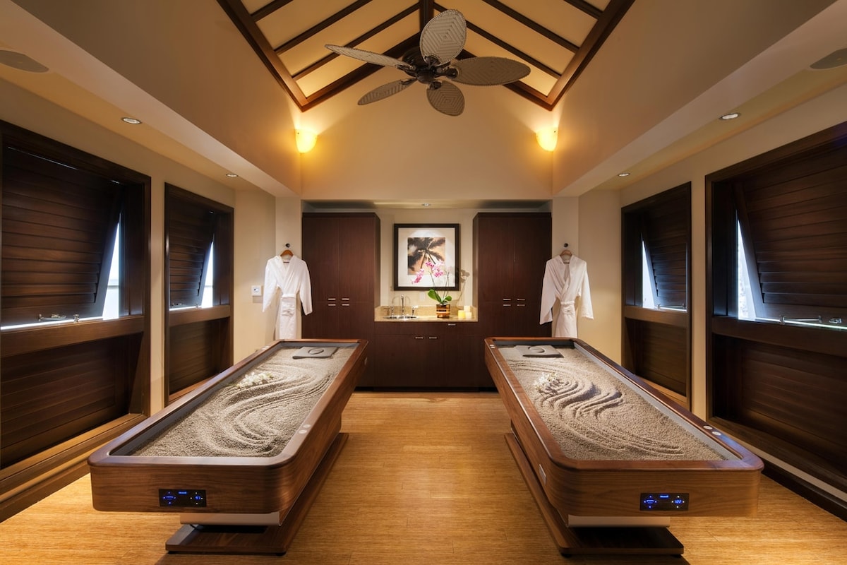 A serene spa room features two wooden treatment tables with smooth sand surfaces. The room is illuminated by soft lighting, and two robes hang on nearby hooks. Large windows with wooden blinds allow for natural light, enhancing a soothing environment.