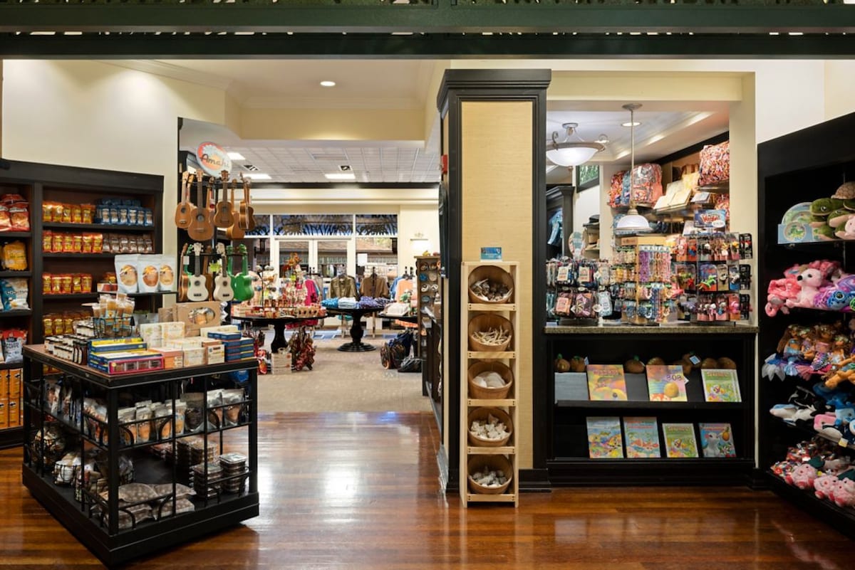 An inviting store interior is displayed, featuring wooden flooring and warm lighting. Shelves are filled with snacks, souvenirs, and toys, while an area in the background is set up with tables and additional merchandise. Soft plush toys and colorful gift items are prominent.