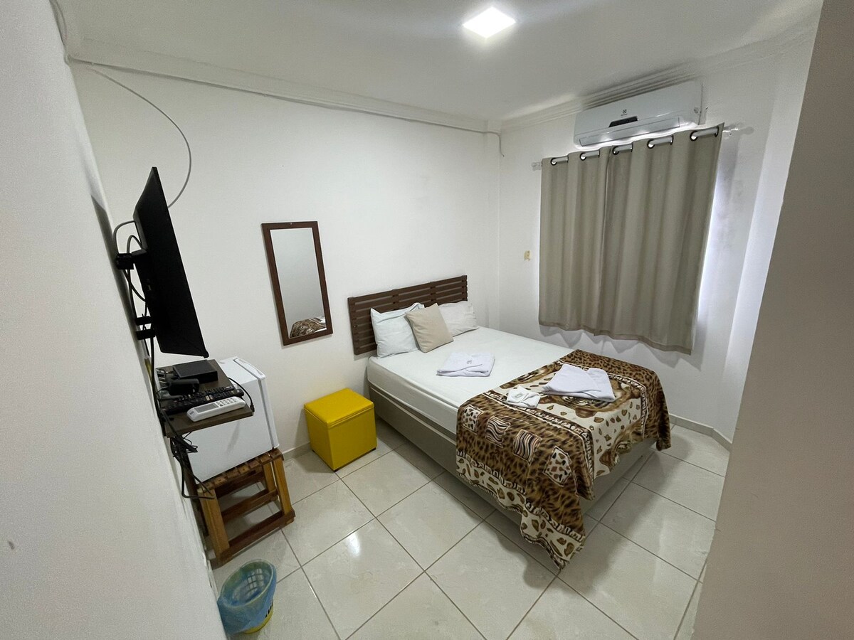 A cozy bedroom features a spacious bed with a patterned blanket and neatly arranged pillows. A large mirror is mounted on the wall, while a smart TV is positioned on a stand opposite the bed. Soft, neutral curtains frame the window, allowing light to filter in.