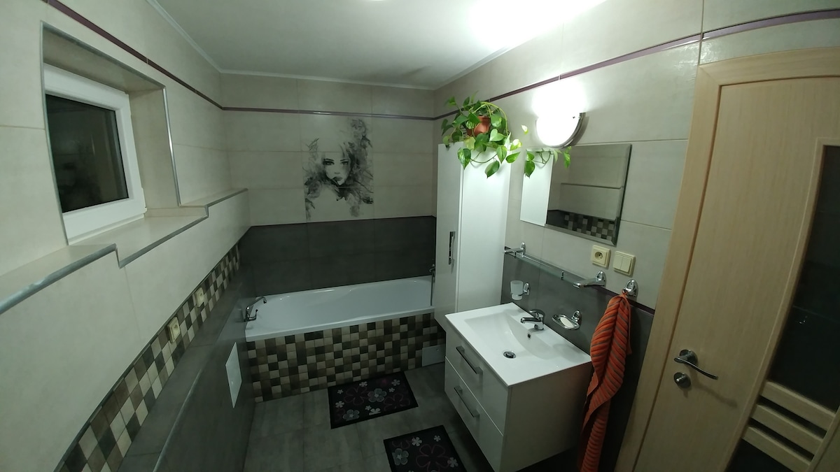 A modern bathroom is equipped with a bathtub, a vanity with sink, and a large mirror. Decorative plants are arranged above the mirror. The space is illuminated by ceiling lights, with a neutral color scheme enhanced by patterned tiles and a stylish towel draped over the vanity.