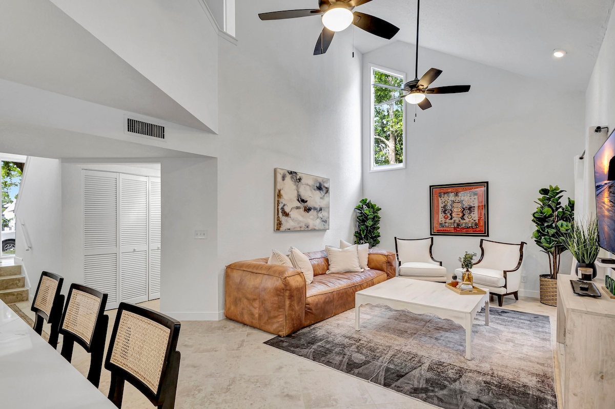 A spacious living area is enhanced by high ceilings and natural light from large windows. A cozy sectional sofa with earth-toned upholstery is centered around a white coffee table, complemented by two accent chairs. Decorative artwork and potted plants add a touch of elegance.