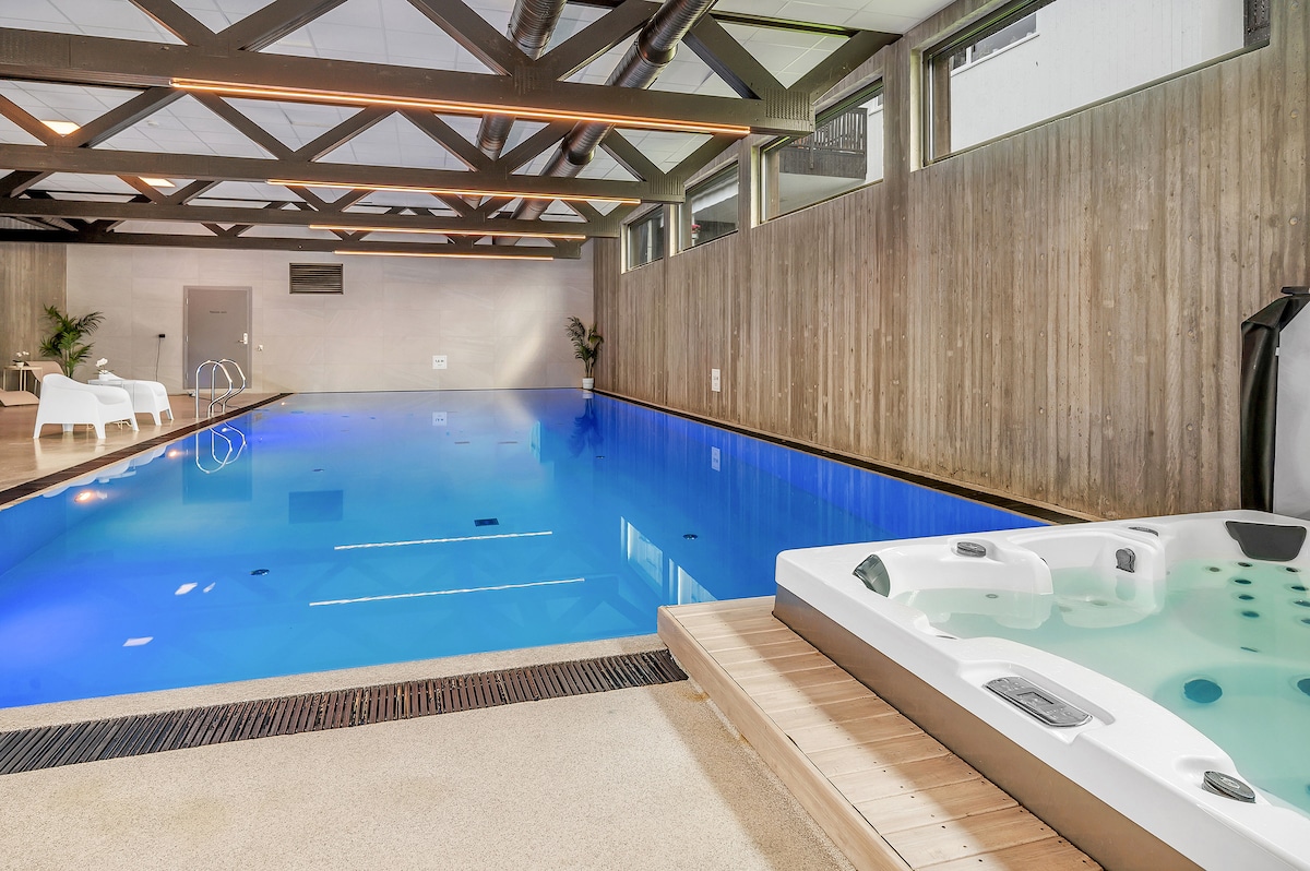 An indoor pool area features a spacious pool with clear blue water and sleek tile edges. Adjacent to the pool, a large hot tub is situated on a wooden deck. Tall windows allow natural light to fill the space, highlighting the wooden beams above.