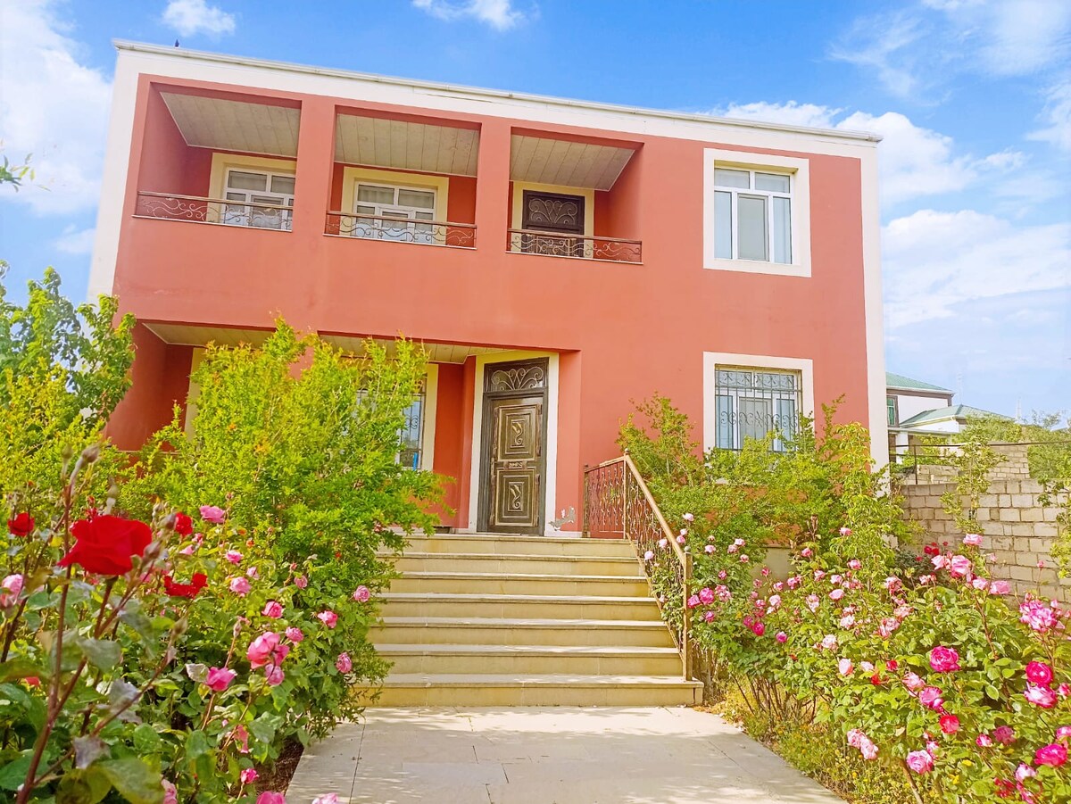 A charming house with a coral-colored facade stands surrounded by lush greenery and vibrant rose bushes. A set of stone steps leads to an ornate front door, complemented by a balcony above. The bright sky adds to the inviting outdoor space.