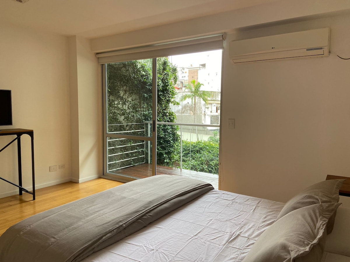 A spacious bedroom is featured, showcasing a large bed with a soft, neutral-colored duvet. Natural light enters through a sliding glass door, which leads to a private balcony with greenery visible outside. A minimalist desk is positioned nearby, and an air conditioning unit is mounted above.