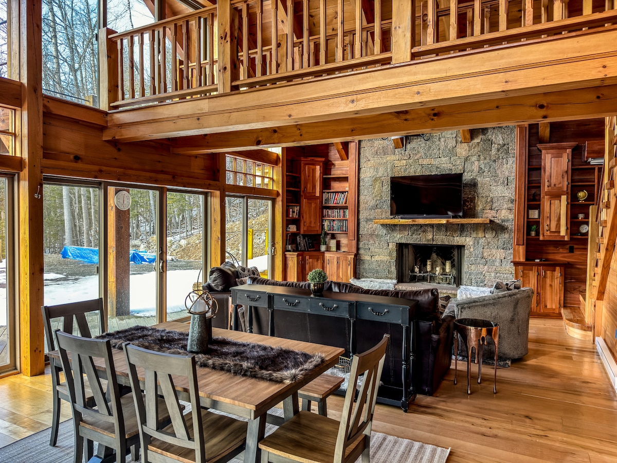 A spacious living area features large windows that allow natural light to fill the space, with views of the outdoors. A stone fireplace serves as a focal point, complemented by comfortable seating and a wooden dining table surrounded by chairs, creating an inviting atmosphere.