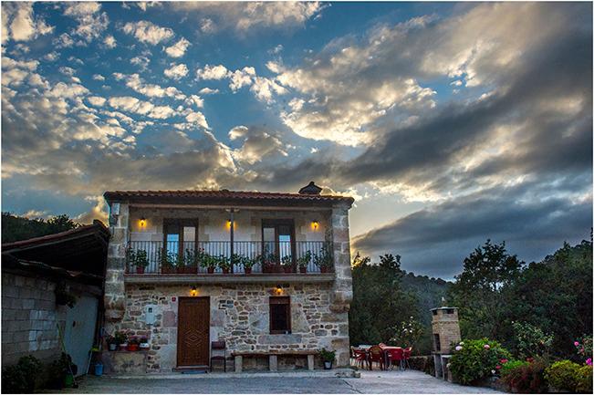A rustic stone exterior is highlighted by multiple windows, adorned with flower boxes. The structure features a classic tile roof and is surrounded by greenery, contributing to a calm ambiance. Dark clouds mix with patches of blue in the evening sky, enhancing the serene setting.