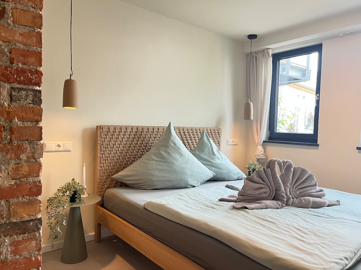 A minimalist bedroom features a spacious bed adorned with two large cushions and a neatly arranged towel sculpture. Soft lighting is provided by pendant lamps, while a window offers natural light, complemented by a small side table with greenery.