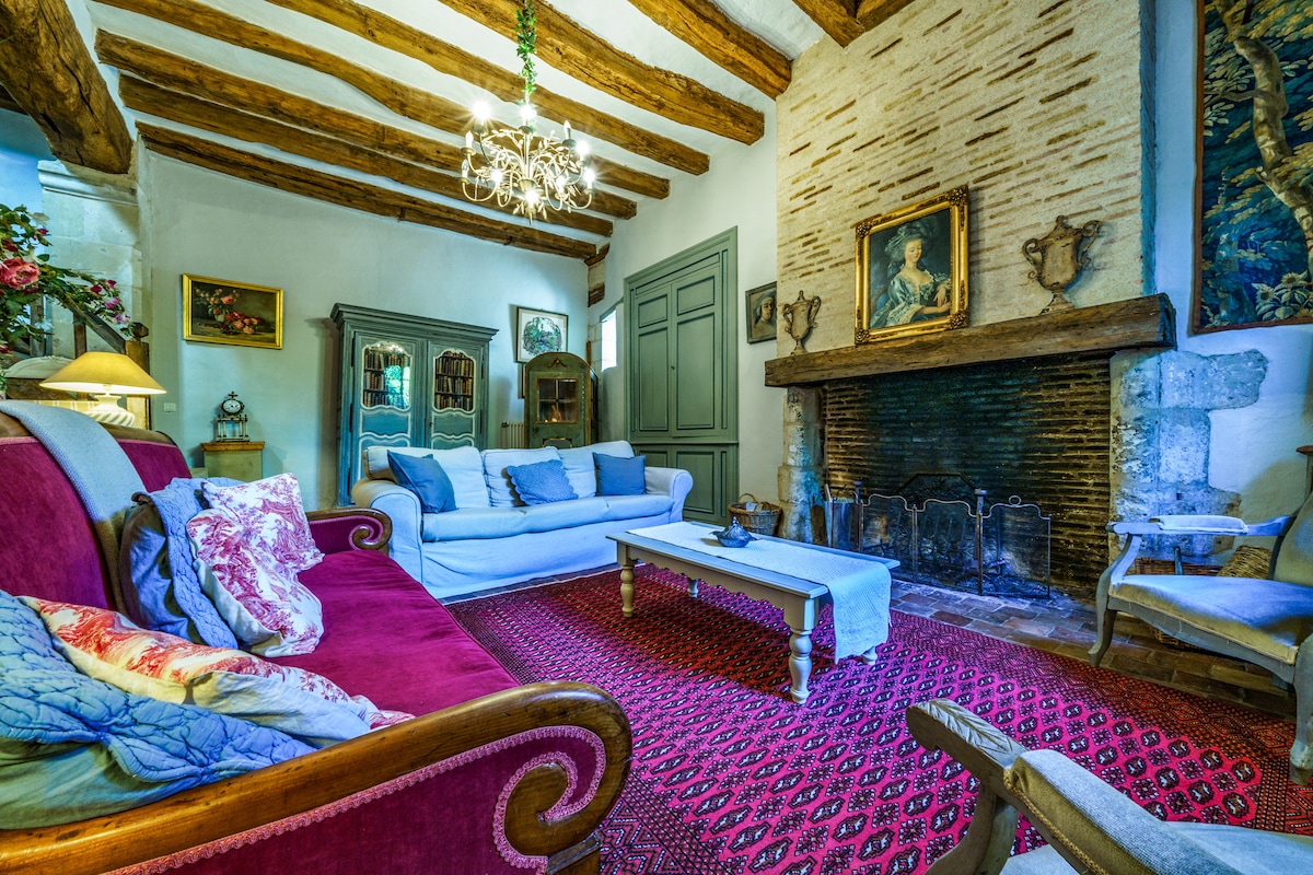 A cozy living room is highlighted by exposed wooden beams and a stone fireplace framed by decorative accessories. A red patterned rug covers the floor, while a mix of vintage and modern furniture, including a white sofa and a red settee, invites relaxation.