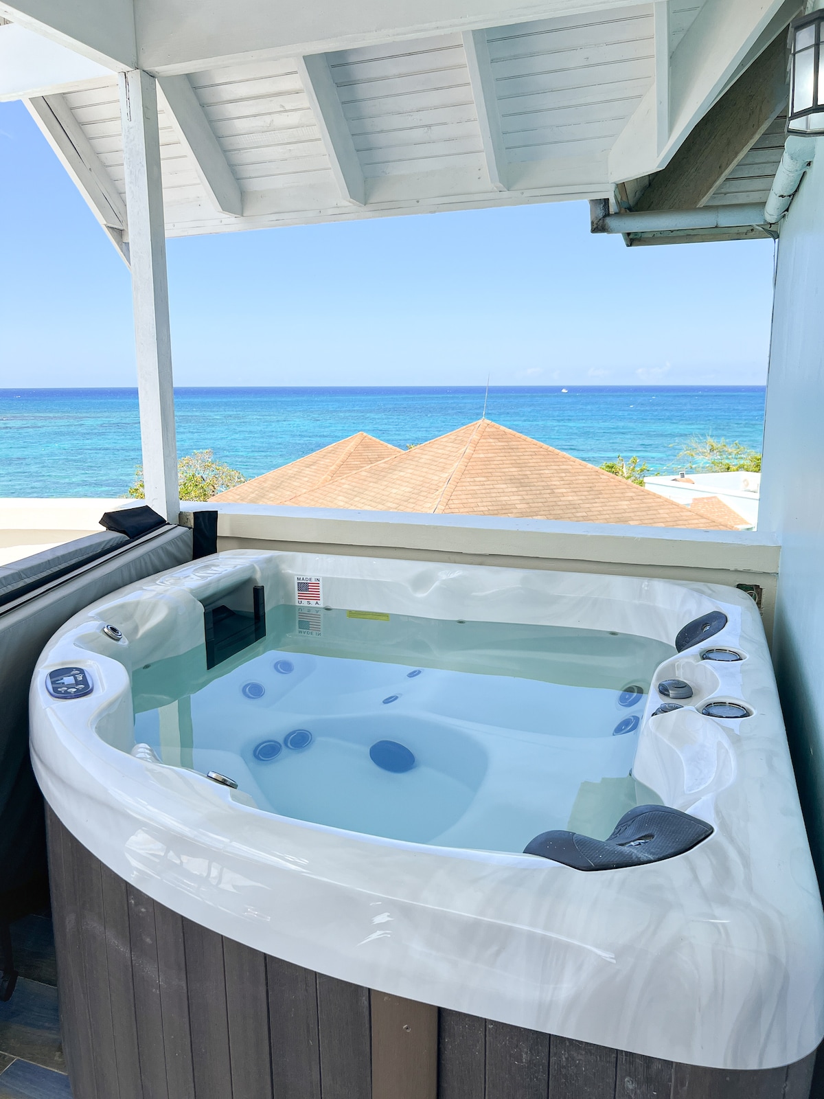 A hot tub is situated on a private balcony, offering views of the Caribbean Sea. Clear water is visible within the tub, surrounded by a wooden deck. The scene includes a light blue sky and ocean waves in the background, creating a serene environment.