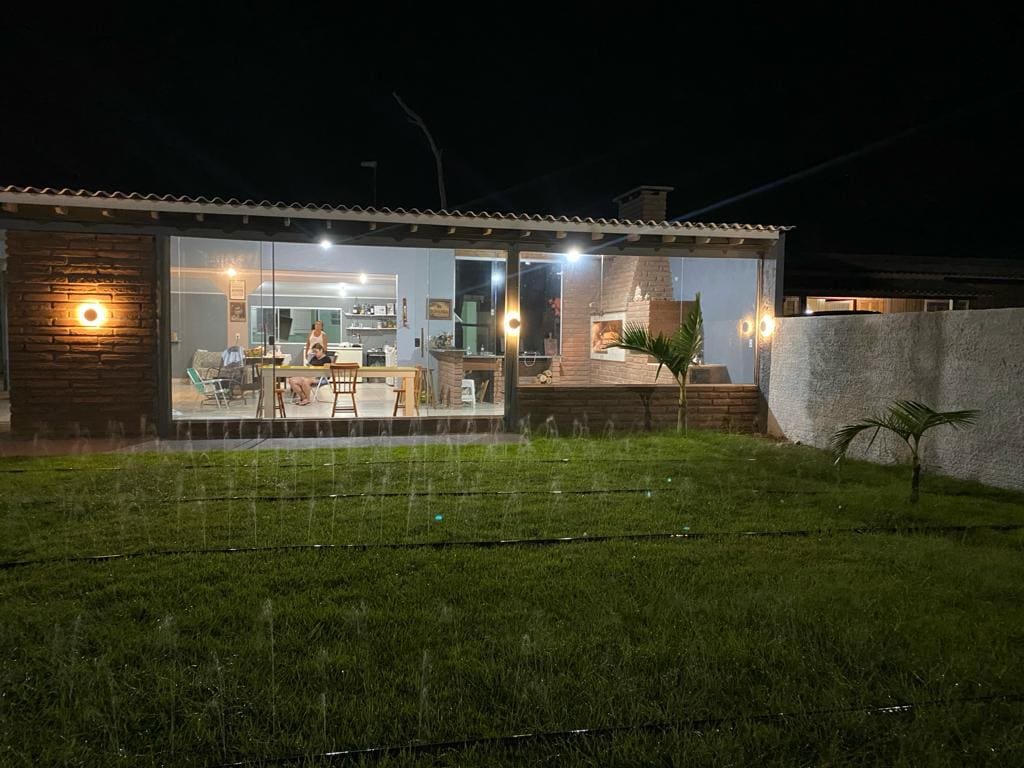 A well-lit, modern house is visible at night, featuring large glass doors that provide a view of the dining area and kitchen. A green lawn with sprinklers running adds freshness to the outdoor space while palm plants are positioned near the house.