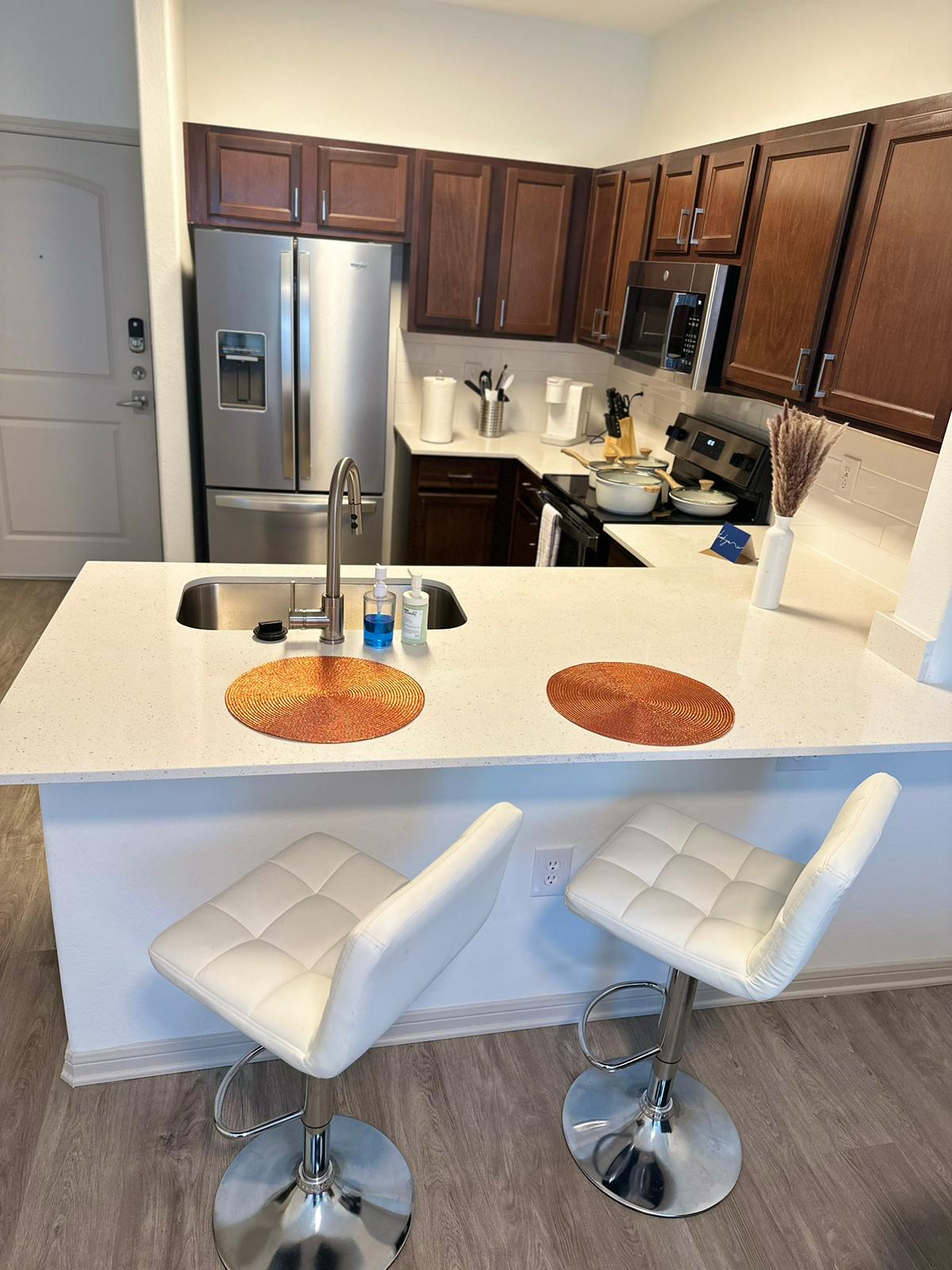 The kitchen features modern wooden cabinetry and stainless-steel appliances. A wide island with a white countertop is complemented by two sleek, white barstools. Bottled items are arranged on the counter, and essentials are organized in the background.